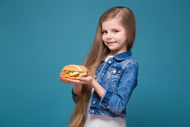 Niña bonita en chaqueta de jean con largo cabello castaño sostener una hamburguesa