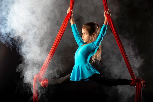 Niña bonita concentrada en un traje de gimnasia azul hace una división en el aire aferrándose a cintas rojas aireadas rodeadas de humo sobre un negro
