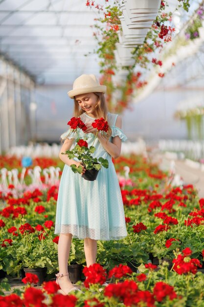 Una niña bonita está de pie en un invernadero y sostiene una olla de flores en sus manos el niño está usando un sombrero