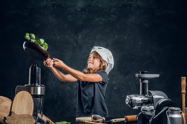 Una niña bonita está tirando verduras en la sartén en un estudio fotográfico oscuro.