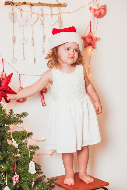 Niña bonita con un gorro de Santa Claus cuelga una estrella en la parte superior del árbol de Navidad