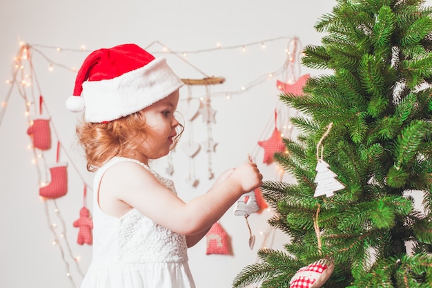 Niña bonita con un gorro de Santa Claus está decorando el árbol de Navidad