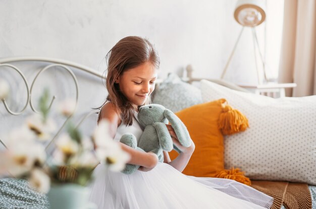 Una niña bonita con un hermoso vestido blanco se sienta en una cama en una habitación luminosa con una liebre de juguete