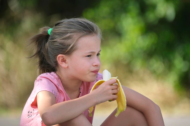 Niña bonita joven comiendo sabrosos bocadillos de plátano maduro al aire libre en el día de verano