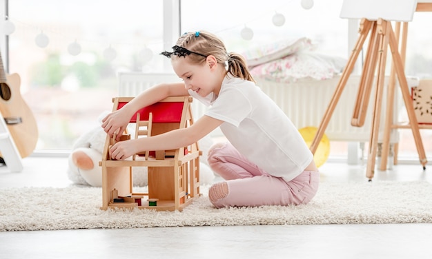 Niña bonita juega con casa de muñecas de madera en la habitación de los niños