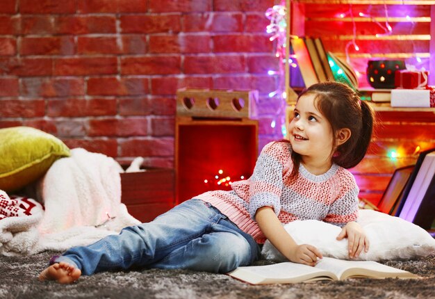 Niña bonita con libro en habitación decorada de Navidad