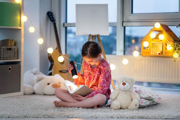 Niña bonita con linterna y libro en el suelo en la habitación crepuscular