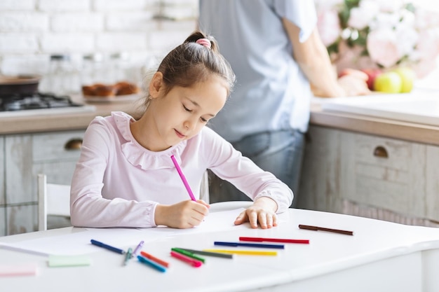 Niña bonita ocupada con el dibujo mientras su madre cocina la comida en la cocina, primer plano