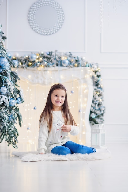 Niña bonita sentada cerca de árbol de Navidad en el interior.