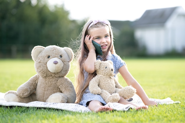 Niña bonita sentada en el parque de verano sobre la hierba verde junto con su osito de peluche hablando por teléfono móvil sonriendo felizmente al aire libre en verano.