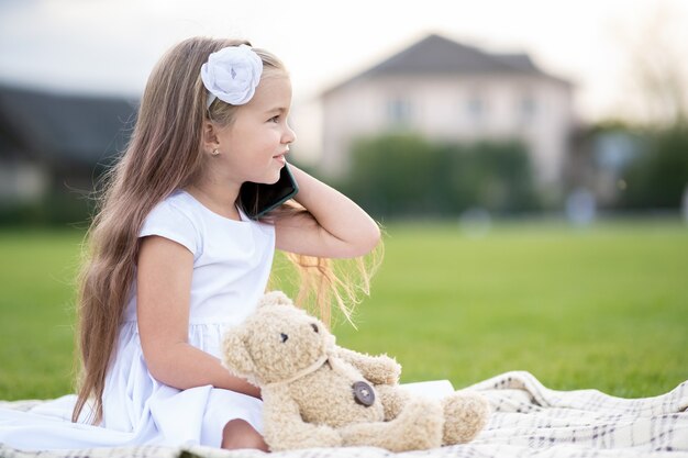 Niña bonita sentada en el parque de verano sobre la hierba verde con su osito de peluche hablando por teléfono móvil al aire libre en verano.