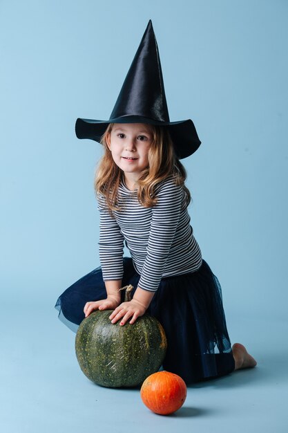 Niña bonita con sombrero de bruja jugando con calabaza verde, apoyándose en él. Está vestida para Halloween. Sobre fondo azul.