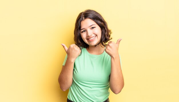 Niña bonita sonriendo con alegría y luciendo feliz, sintiéndose despreocupada y positiva con ambos pulgares hacia arriba