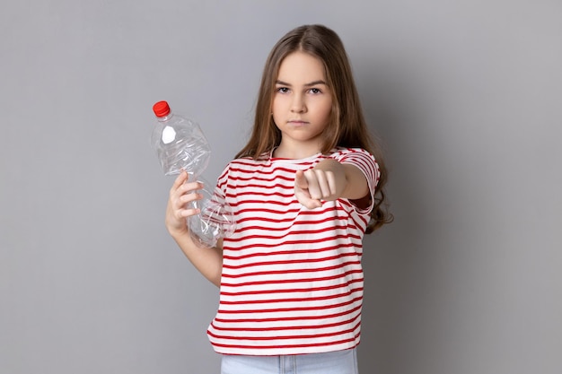 Niña bonita sosteniendo una botella de plástico vacía mirando y señalando con el dedo a la cámara