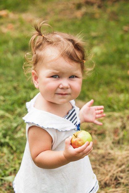 Niña bonita sosteniendo una manzana en la mano, de pie bajo el manzano