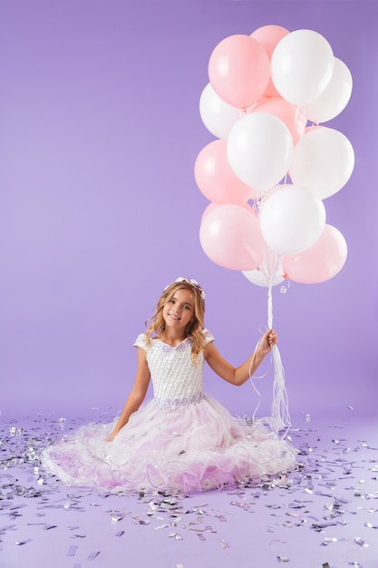 Niña bonita vestida con traje de princesa sentada aislada sobre pared violeta, sosteniendo un montón de globos