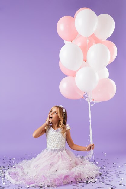 Niña bonita vestida con traje de princesa sentada aislada sobre pared violeta, sosteniendo un montón de globos