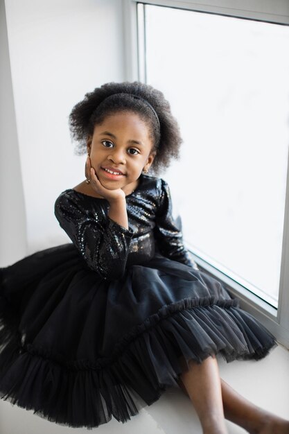 Niña bonita con vestido negro sentada cerca de la ventana