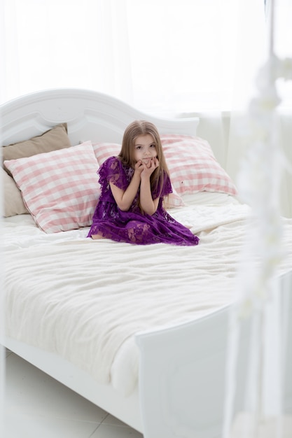 Niña bonita en vestido de princesa púrpura en cama blanca con cojín