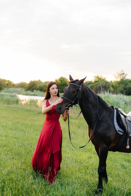 Una niña bonita con un vestido rojo posa en un rancho con un semental de pura sangre al atardecer. Amar y cuidar a los animales.