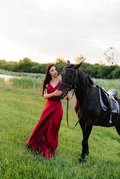 Una niña bonita con un vestido rojo posa en un rancho con un semental de pura sangre al atardecer. Amar y cuidar a los animales.