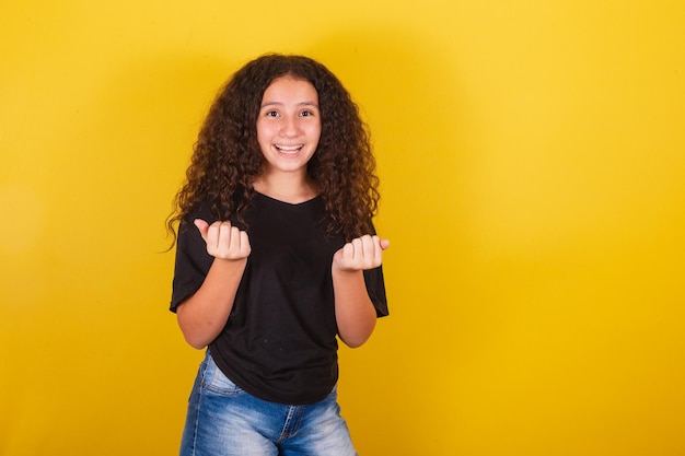 Niña brasileña latinoamericana de cabello afro fondo amarillo sonriendo indicando ven bienvenido ven con manos y dedos ven aquí expresión