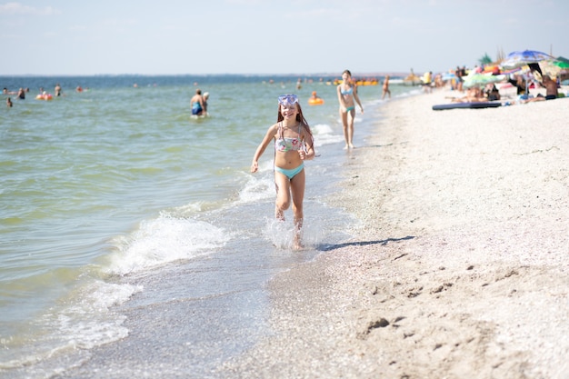 una niña bronceada con el pelo largo en un traje de baño azul corre alegremente a lo largo de la orilla arenosa cerca del Mar Negro