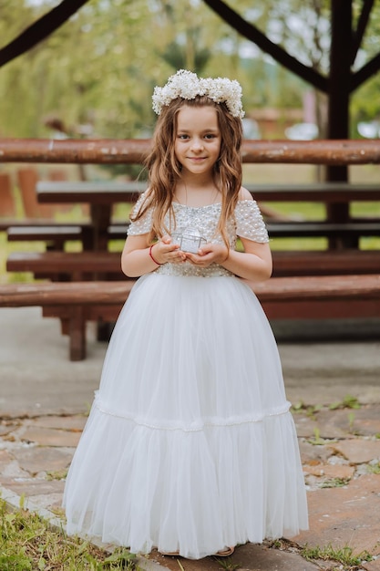 una niña de cabello claro con flores en la cabeza en un vestido blanco sosteniendo anillos de bodas para la ceremonia de boda boda de primavera