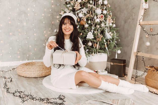 Una niña con cabello largo oscuro y un gorro de punto blanco se sienta cerca de un árbol de Navidad y tiene un regalo en sus manos.