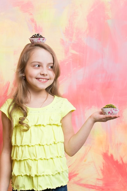 Niña con cabello largo y rubio sosteniendo pasteles Niña sonriendo con pastelitos en la mano y en la cabeza