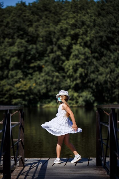 Una niña de cabello largo con un vestido blanco se encuentra en la orilla del río. Camina por el lago en un día soleado de verano