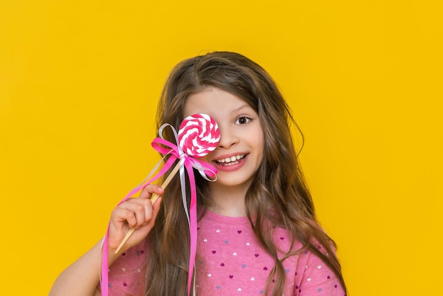 Una niña con un cabello muy hermoso y un vestido rosa sostiene una piruleta en sus manos Una niña muy alegre y alegre con un gran caramelo en un palo en un fondo aislado