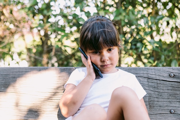 Niña de cabello negro, vestida con una camiseta blanca, sentada en un banco en un parque hablando por teléfono móvil, con un gesto serio.