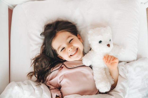 Una niña de cabello oscuro está durmiendo en la cama abrazando a un oso de peluche debajo de una manta blanca. El niño está en la cama por la mañana.