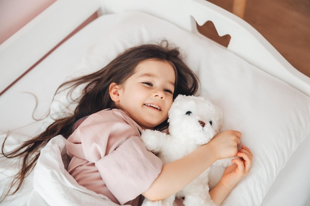 Una niña de cabello oscuro está durmiendo en la cama abrazando a un oso de peluche debajo de una manta blanca. El niño está en la cama por la mañana.