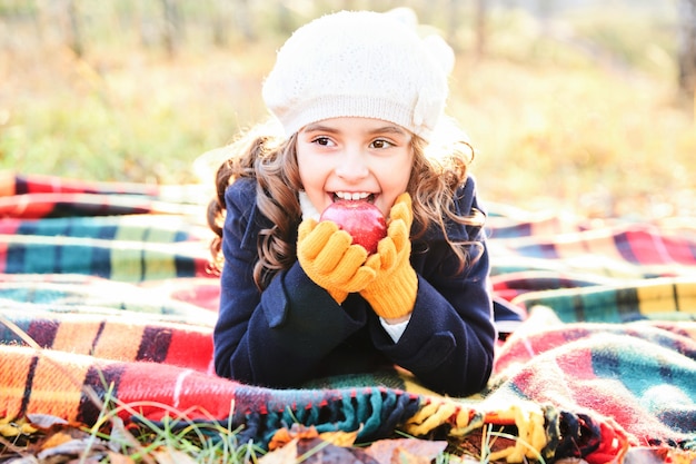 Niña de cabello oscuro sonriendo acostado sobre una manta con una manzana en sus manos en el parque en otoño. Foto de alta calidad