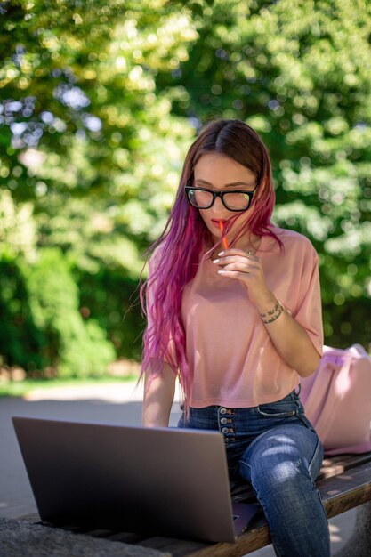 Niña con cabello rosado está estudiando en el parque de la primavera, sentada en el banco de madera y navegando en su computadora portátil