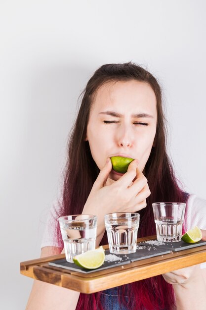 Niña con cabello rosado sostiene una bandeja de madera con chupitos de tequila y come lima