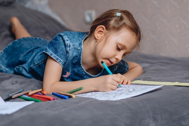 Una niña con cabello rubio se acuesta en la cama y elige el color de un rotulador para colorear la imagen