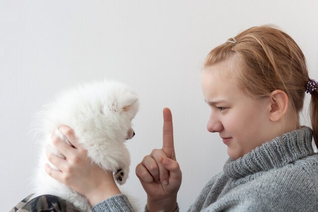 Una niña de cabello rubio con un suéter gris, con rostro severo, sostiene un cachorro de Pomerania blanco y esponjoso en sus brazos y le sacude el dedo índice al cachorro.