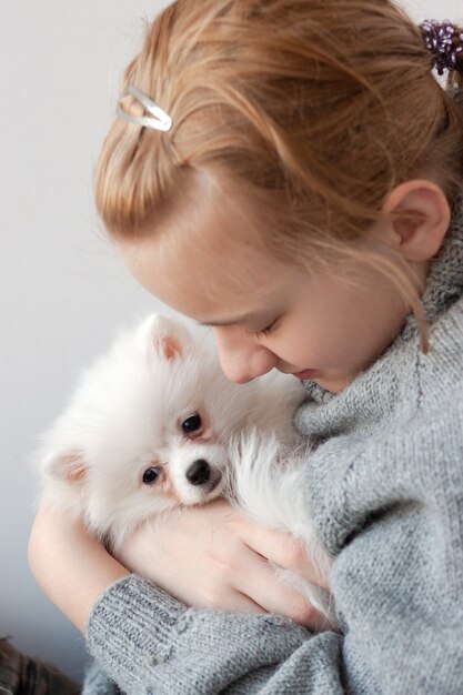 Una niña de cabello rubio con un suéter gris sostiene a un cachorro de Pomerania blanco y esponjoso en sus brazos, abraza al cachorro y abraza al cachorro.