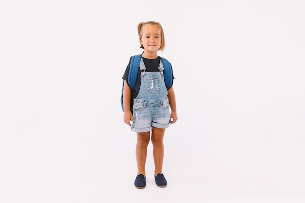Niña con cabello rubio vestida con un peto azul y camiseta, con una mochila lista para volver a la escuela, sobre fondo blanco.