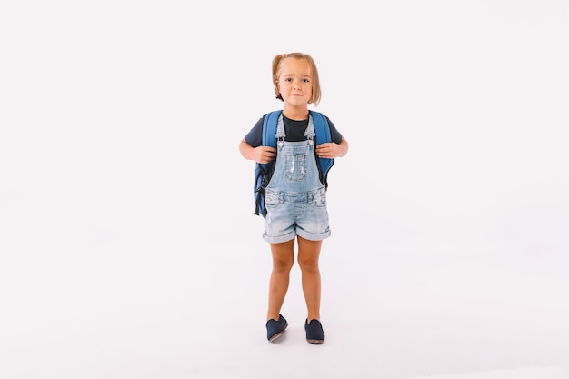 Niña con cabello rubio vestida con un peto azul y camiseta, con una mochila lista para volver a la escuela, sobre fondo blanco.