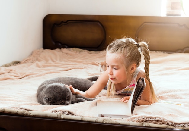 Una niña con cabello rubio yace en una cama en su casa con un gato leyendo un libro. Quédate en casa. Educación a domicilio para niños.