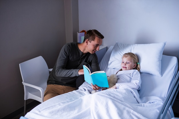 Niña en una cama de hospital leyendo un libro con su padre