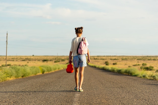 Una niña camina por un camino vacío en la estepa. Tiene un ukelele en las manos y una mochila.