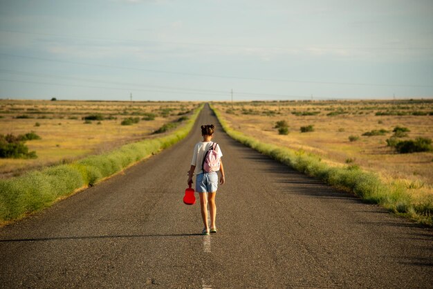 Una niña camina por un camino vacío en la estepa. Tiene un ukelele en las manos y una mochila.