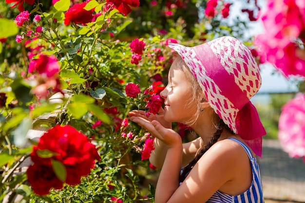 Niña camina en el jardín con rosas en flor. retrocede en un sombrero con una cinta rosa, espacio para texto