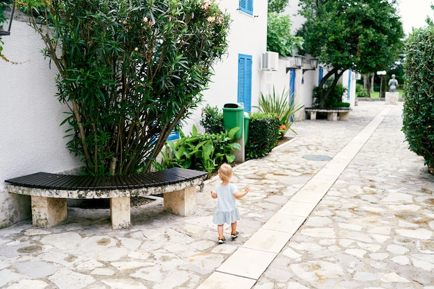 Niña camina por el patio adoquinado cerca de la casa un banco y vista posterior de la vegetación