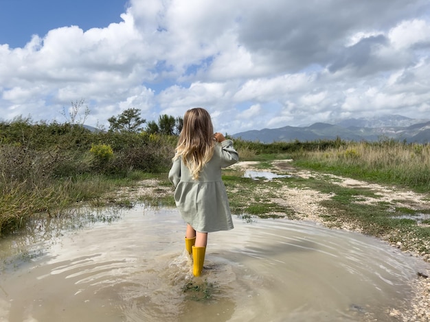 La niña camina a través de un charco hacia un camino rural y mira las montañas